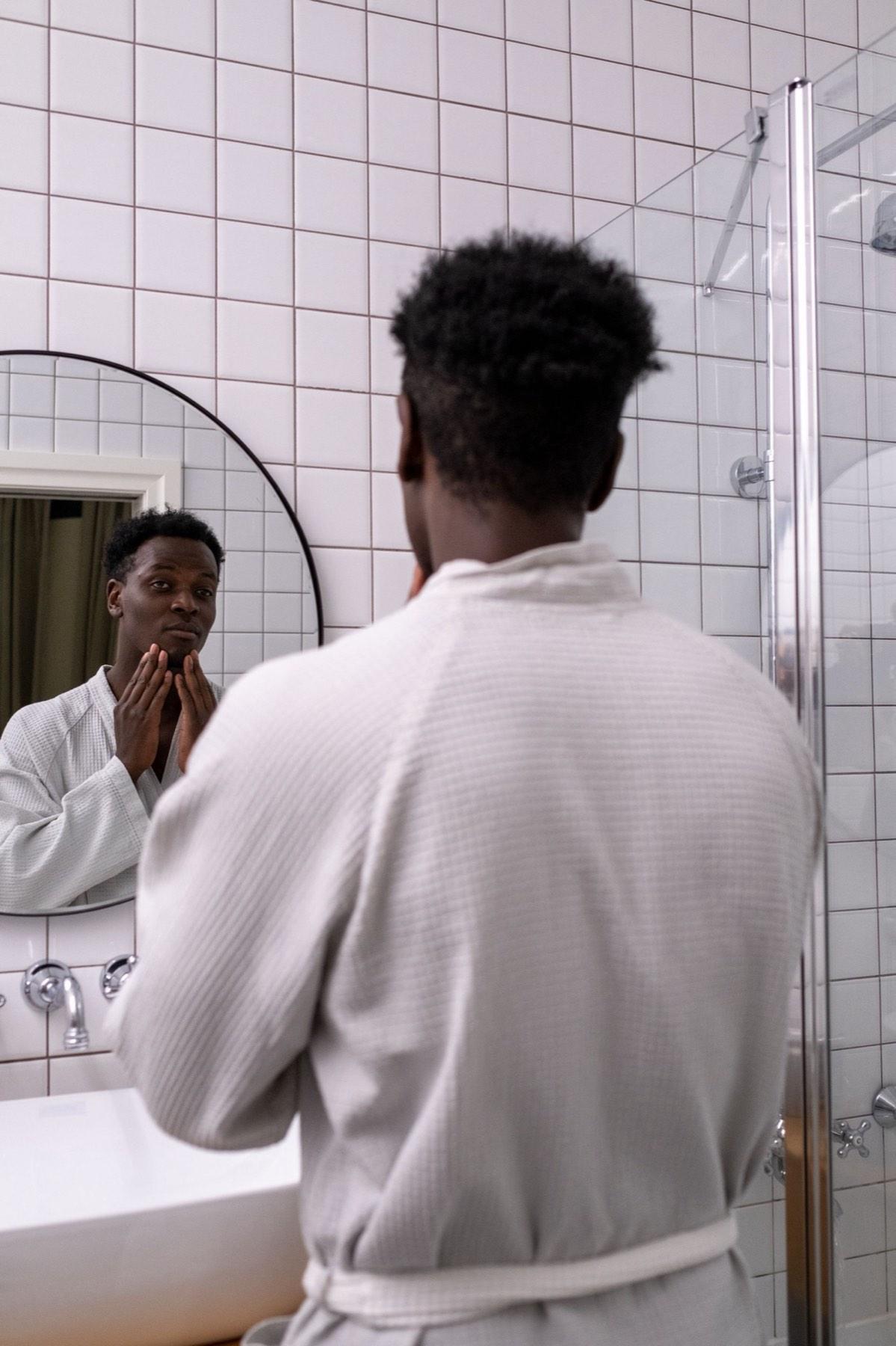 Man in a bathrobe checking his face and hairline in a bathroom mirror