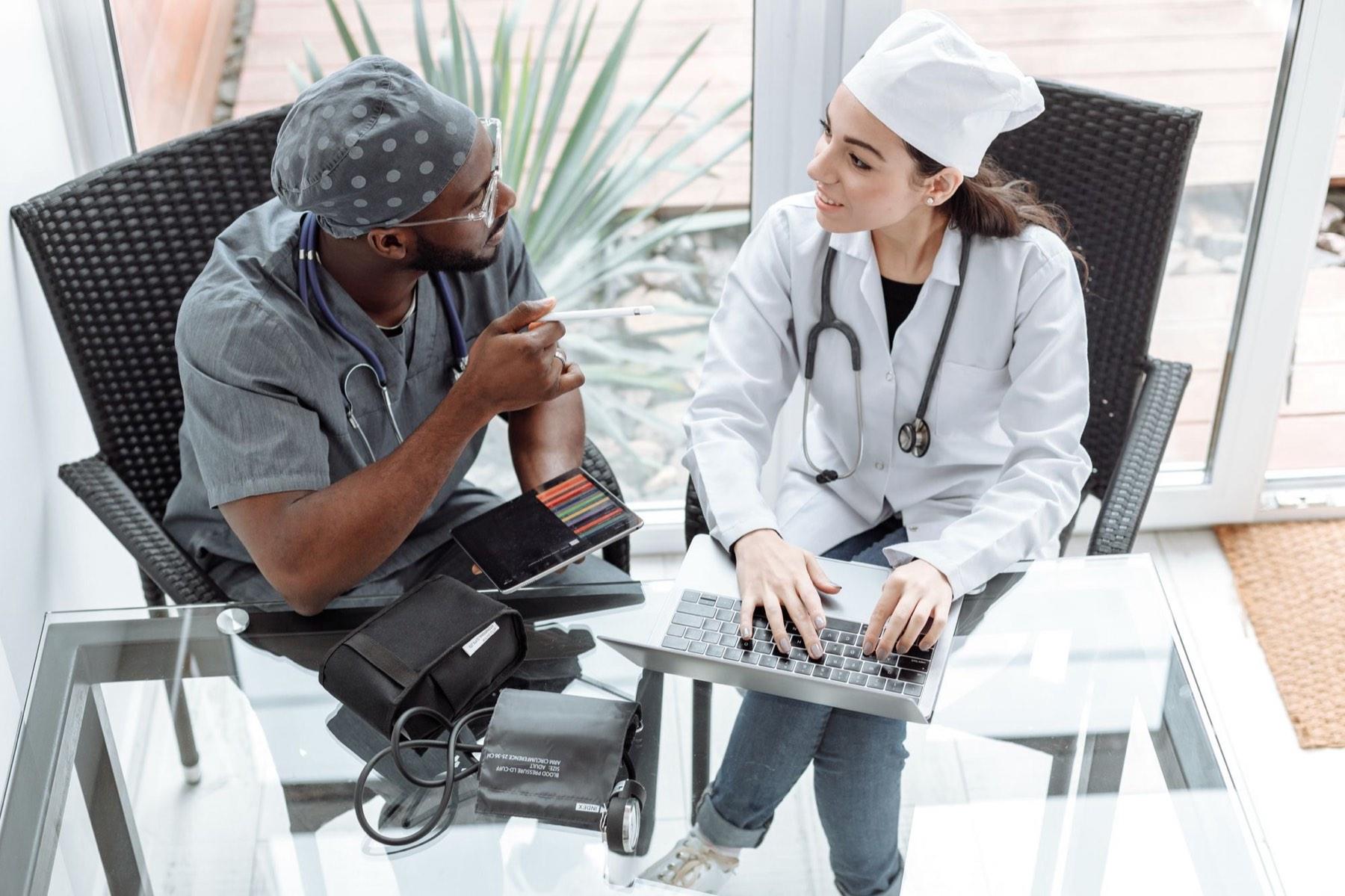 Two clinicians comparing notes over a laptop and tablet
