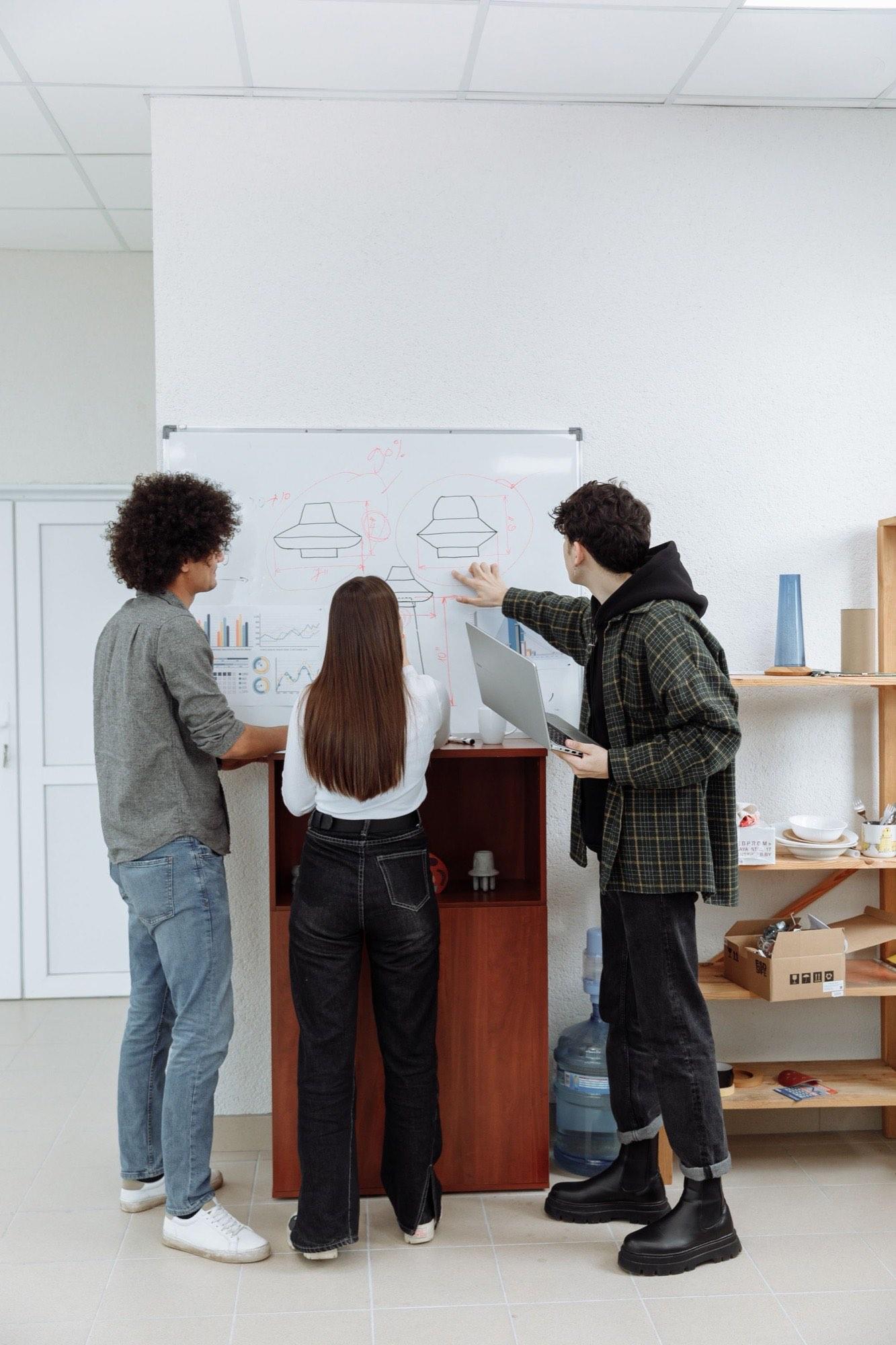 Three teammates sketching product drawings on a whiteboard, one holding a laptop