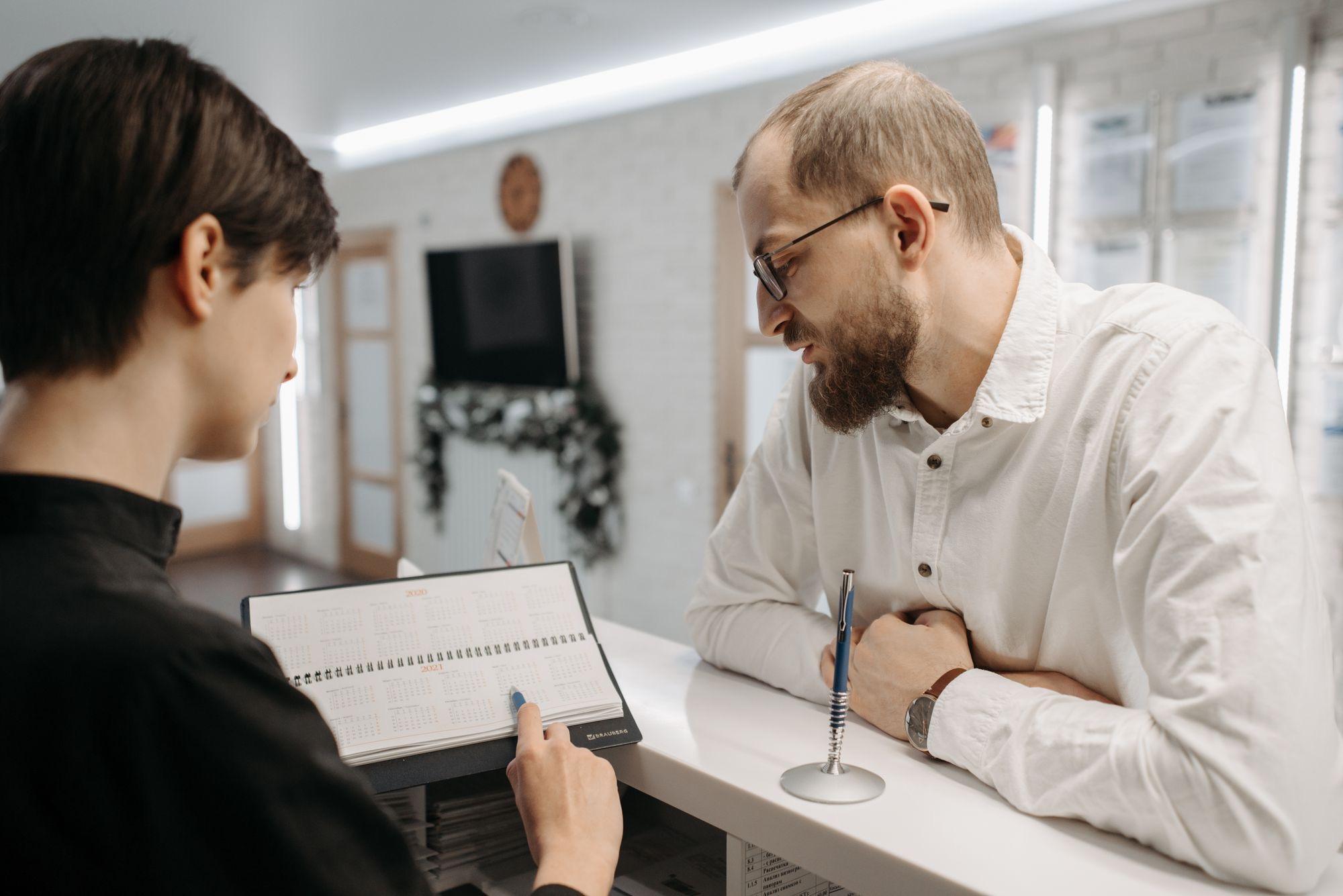 Receptionist pointing to a date in an appointment book while a patient leans on the clinic front desk